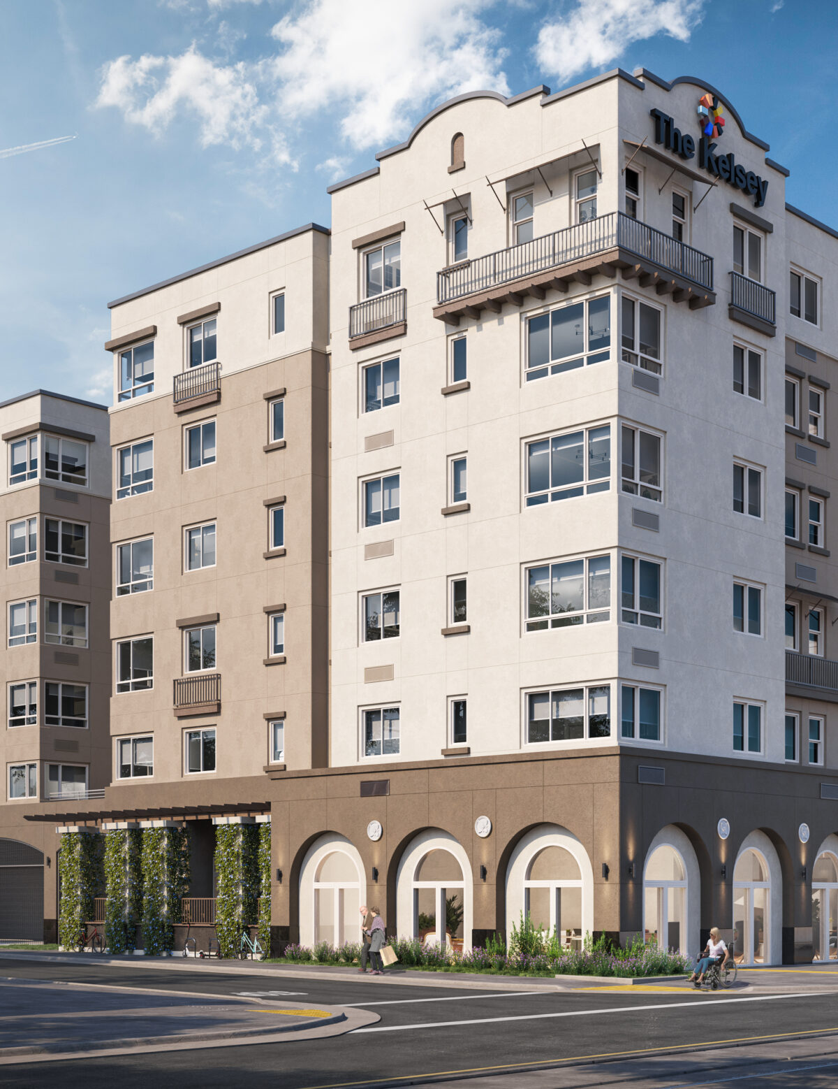 The exterior of The Kelsey Ayer Station, a tan and brown building with windows, small balconies, and arched windows and greenery on the bottom floor. A sign on the top says "The Kelsey" with our brandmark above it. A train is in the bottom right corner about to pass by and people using wheelchairs and walking are on the street.