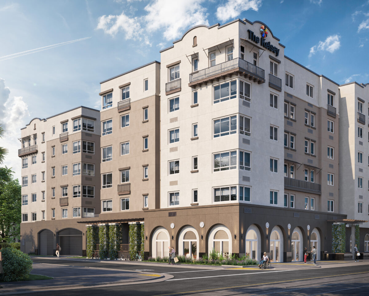 The exterior of The Kelsey Ayer Station, a tan and brown building with windows, small balconies, and arched windows and greenery on the bottom floor. A sign on the top says "The Kelsey" with our brandmark above it. A train is in the bottom right corner about to pass by and people using wheelchairs and walking are on the street.