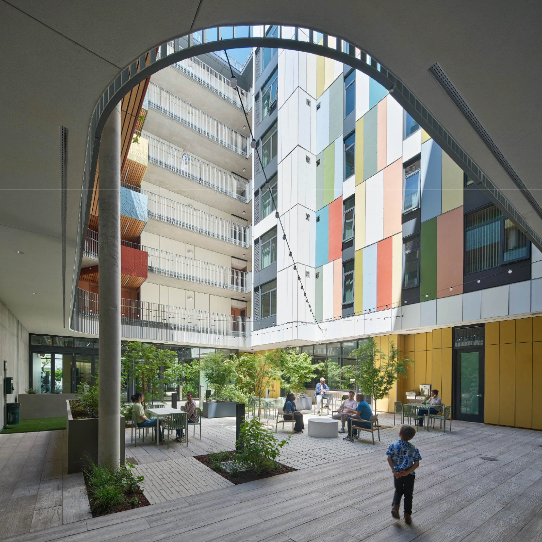 An angled view of the courtyard of The Kelsey Civic Center. Tables and chairs with people sitting at them and trees are visible. Strings of lights hang across the courtyard.