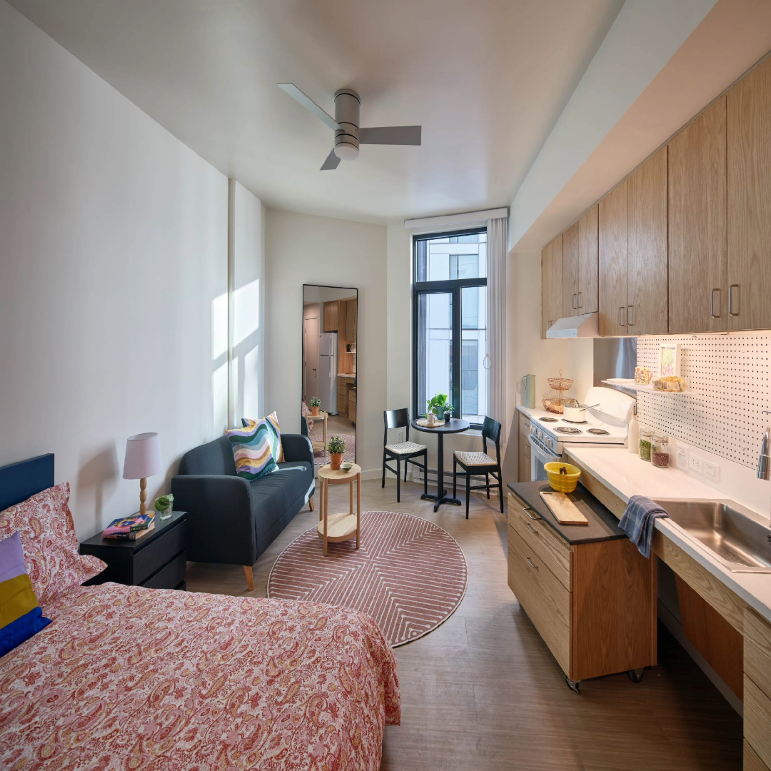 A studio apartment at The Kelsey Civic Center. The room is colorfully decorated with a bed, a nightstand, a couch and a table and chairs. There is a counter wth a sink and a stove.