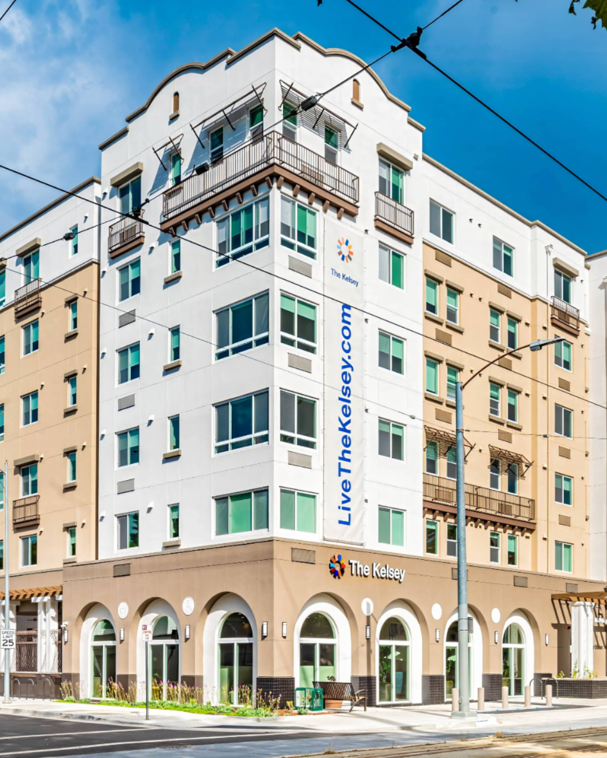 The exterior of The Kelsey Ayer Station, an apartment building with many windows and white and beige paneling. A banner running down the side of the building reads "Livethekelsey.com"