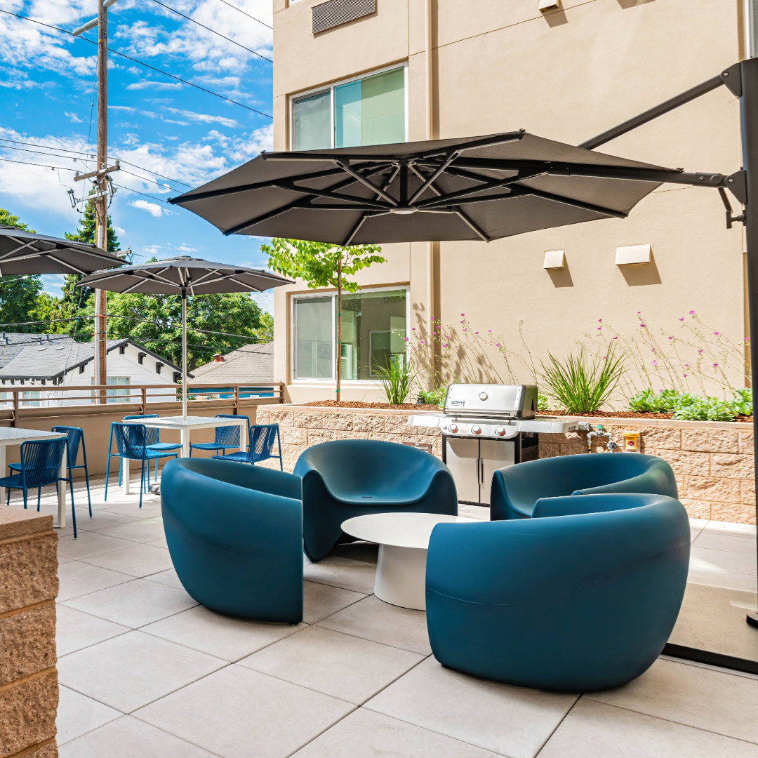 The outdoor patio at The Kelsey Ayer Station. Four large blue chairs around a table with an umbrella are visible. A grill and a white table with four other chairs can be seen in the background.