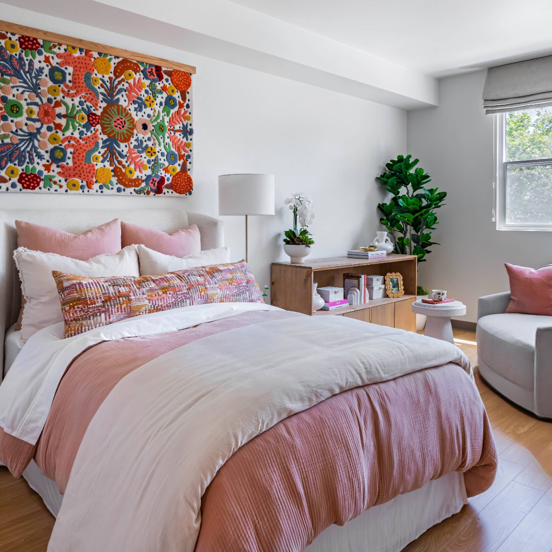 A bedroom with a bed that has pink and white bedding. There is a lamp, a bookshelf, and a plant next to the bed. There is also a gray chair with pink pillows on it near the bed. Artwork is visible on one of the walls.