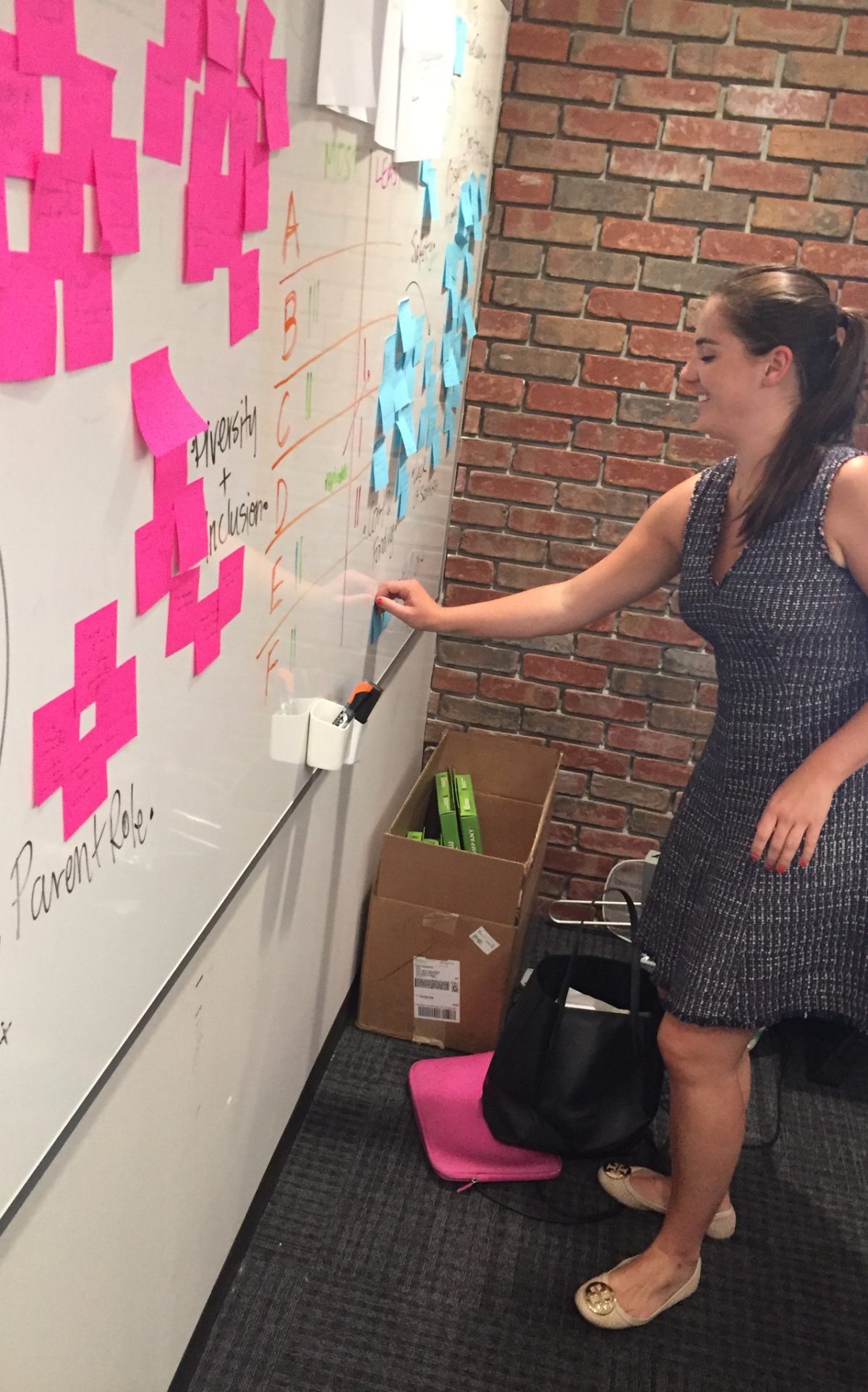 Photo of Lindsay, woman with dark hair pulled back in grey dress, posting pink posted notes on wall.