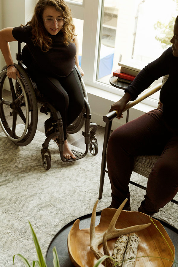 Allie Cannington is pictured in her wheelchair. She is a light skinned person with long dark hair. She is wearing a dark t-shirt and dark pants. She is pictured near a person sitting in a chair.