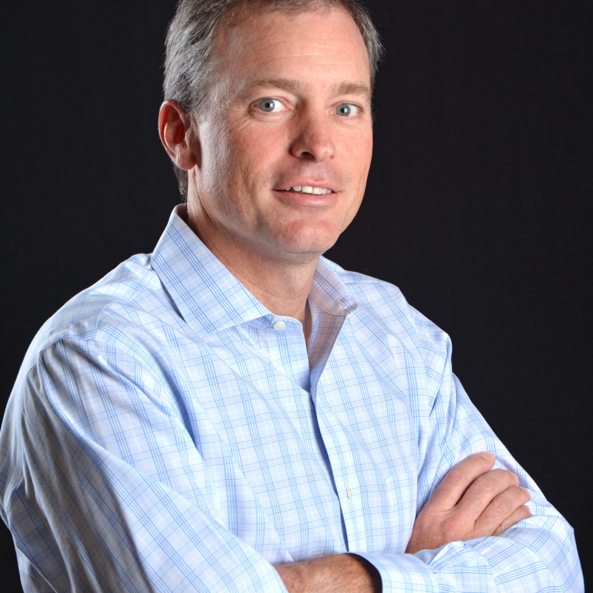 Chris Beda is pictured. He is a light-skinned man with short hair. His arms are crossed. He is wearing a white and blue button down shirt.