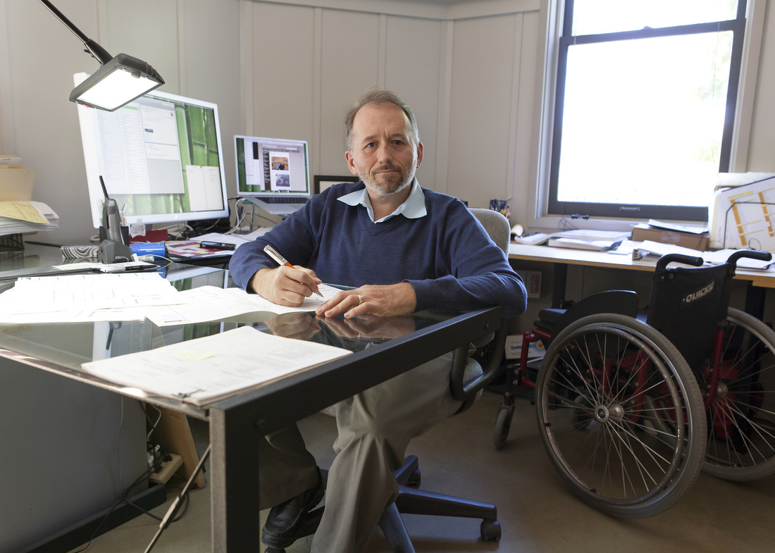 Erick Mikiten, a white man with glasses, wearing a blue sweater, a blue shirt underneath, and gray dress pants is pictured sitting on a chair at his desk. His wheelchair is pictured in the background,