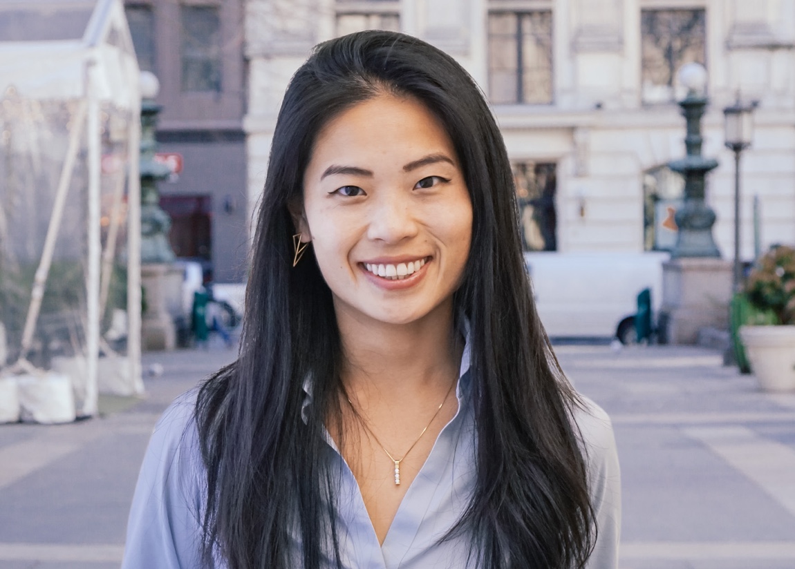 A young asian woman with long, dark hair wearing a necklace and a light purple shirt. She is outdoors. A building is pictured in the background behind her,