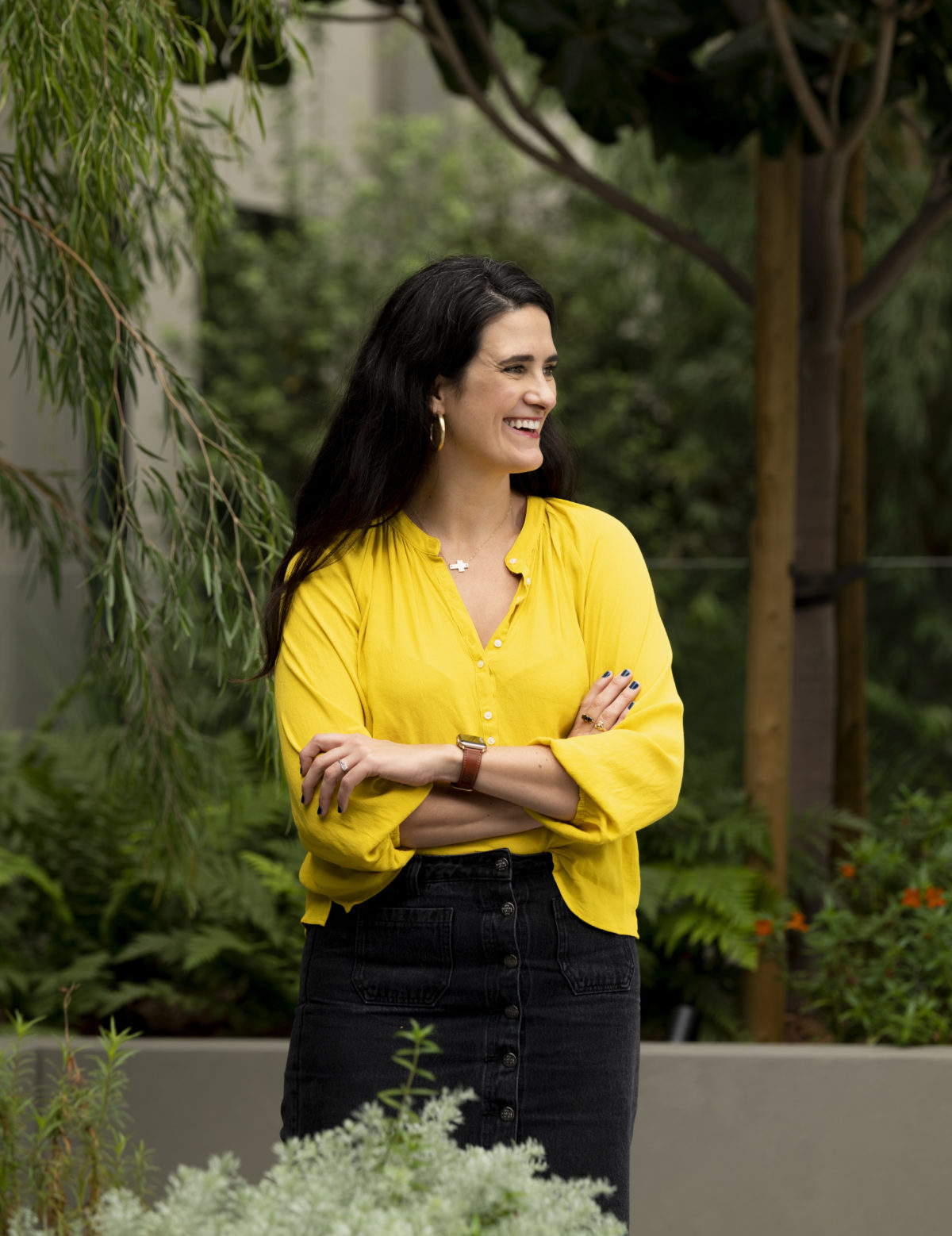 Micaela is white woman in her thirties. She has long brown hair, and is wearing a yellow blouse and black skirt. There is greenery surrounding her.