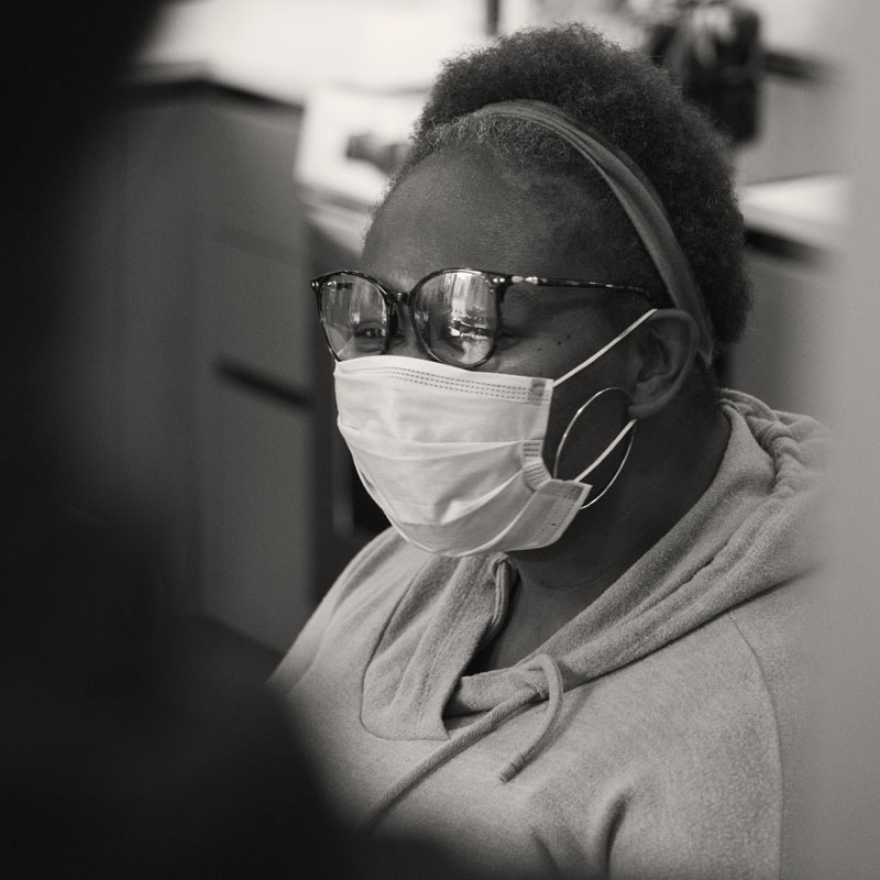 A black and white picture of a dark skinned woman with short, dark hair wearing a mask.