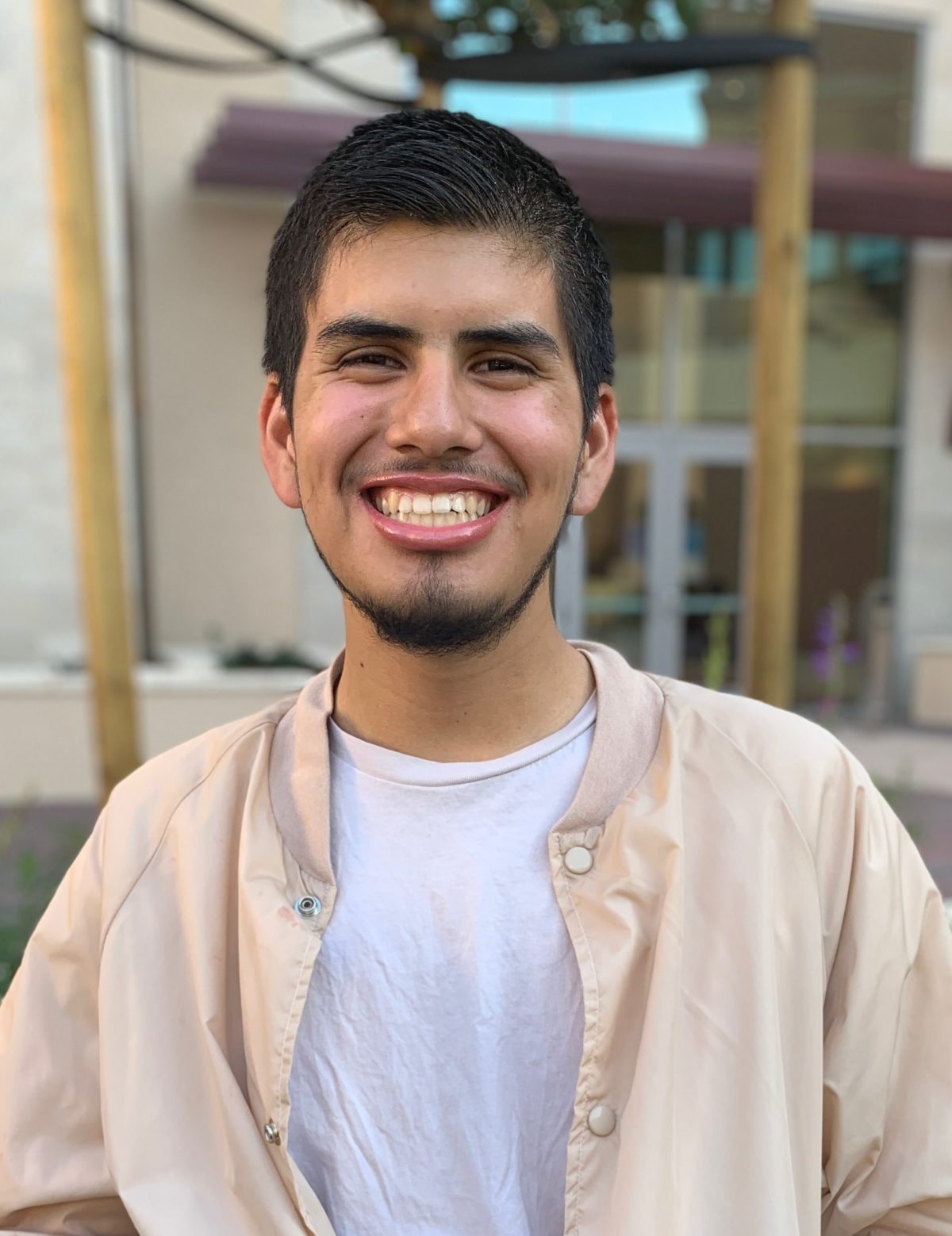 A young Latinx male adult wearing a tan jacket and beige shirt smiles brightly at the camera in front of a residential building.