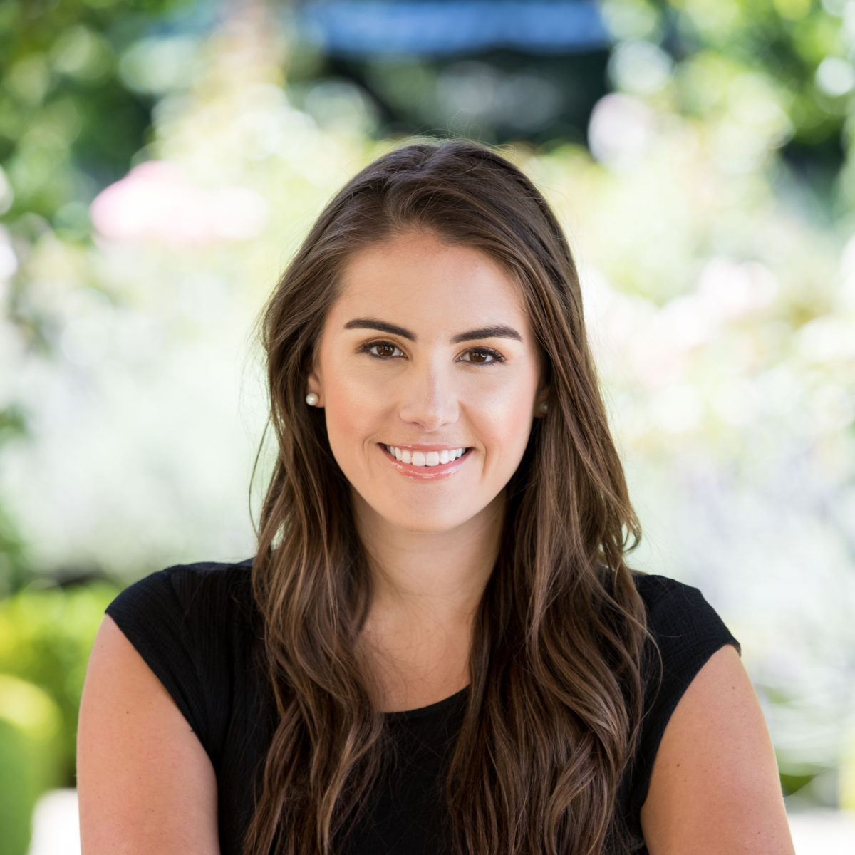 Lindsay Johnson, a light skinned person with long brown hair is pictured against blurred greenery wearing a black outfit. She is smiling.