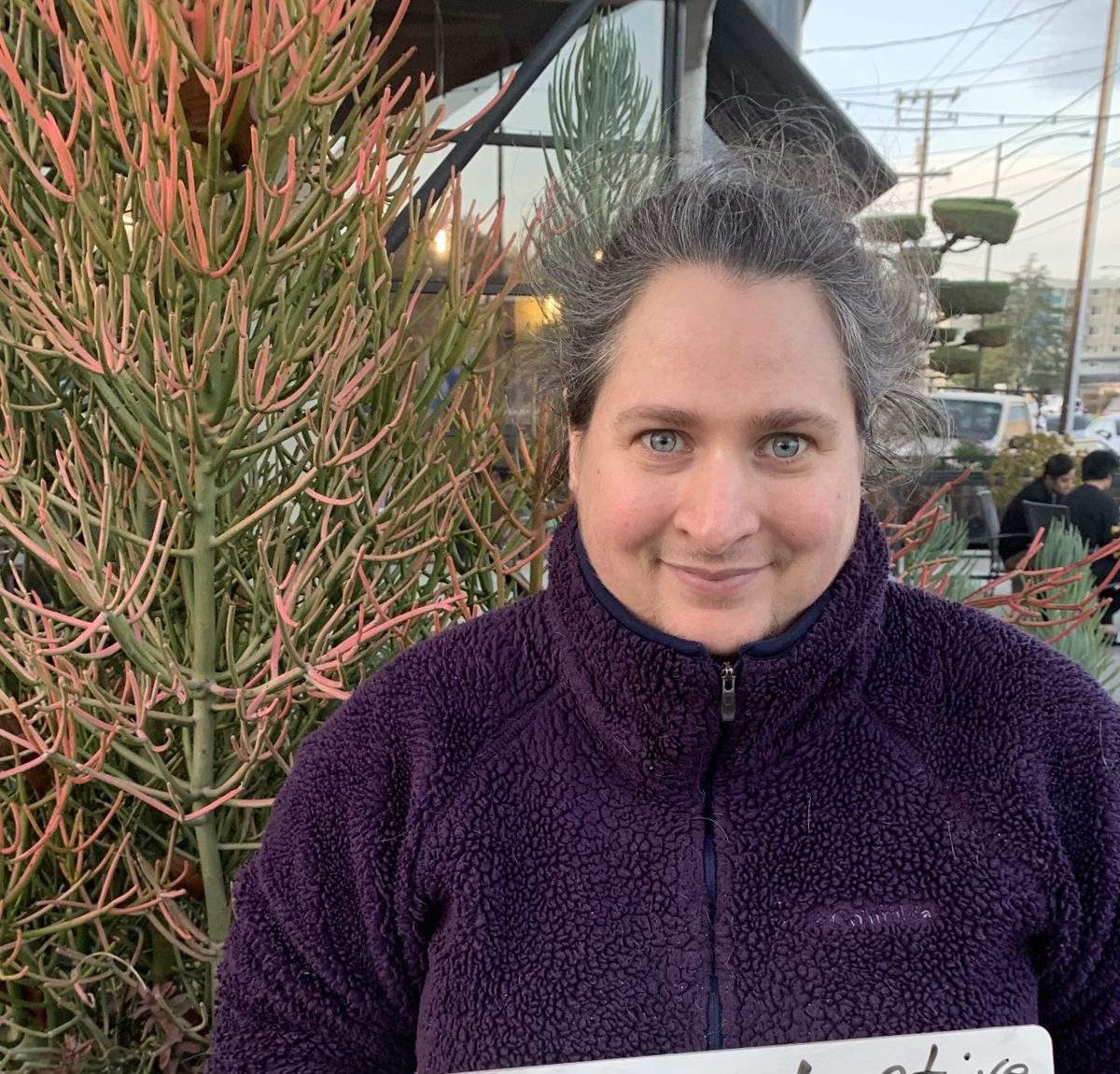 Myra, a young white woman with green eyes, wearing a purple jacket, smiling at the camera with a plant behind her.