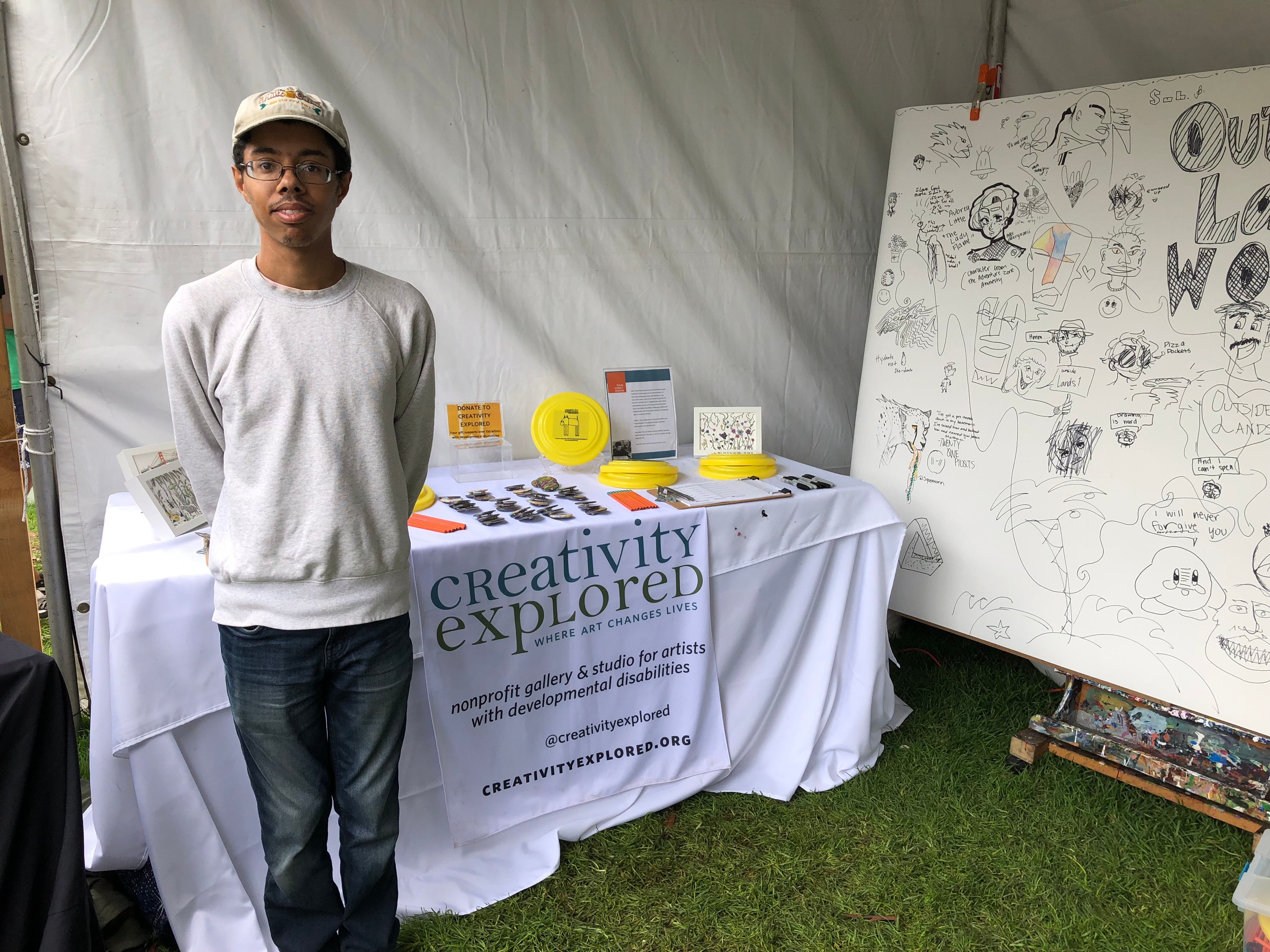 Isaac, black male smiling at camera, wearing glasses and a hat, standing in front of Creativity Explored table at event.