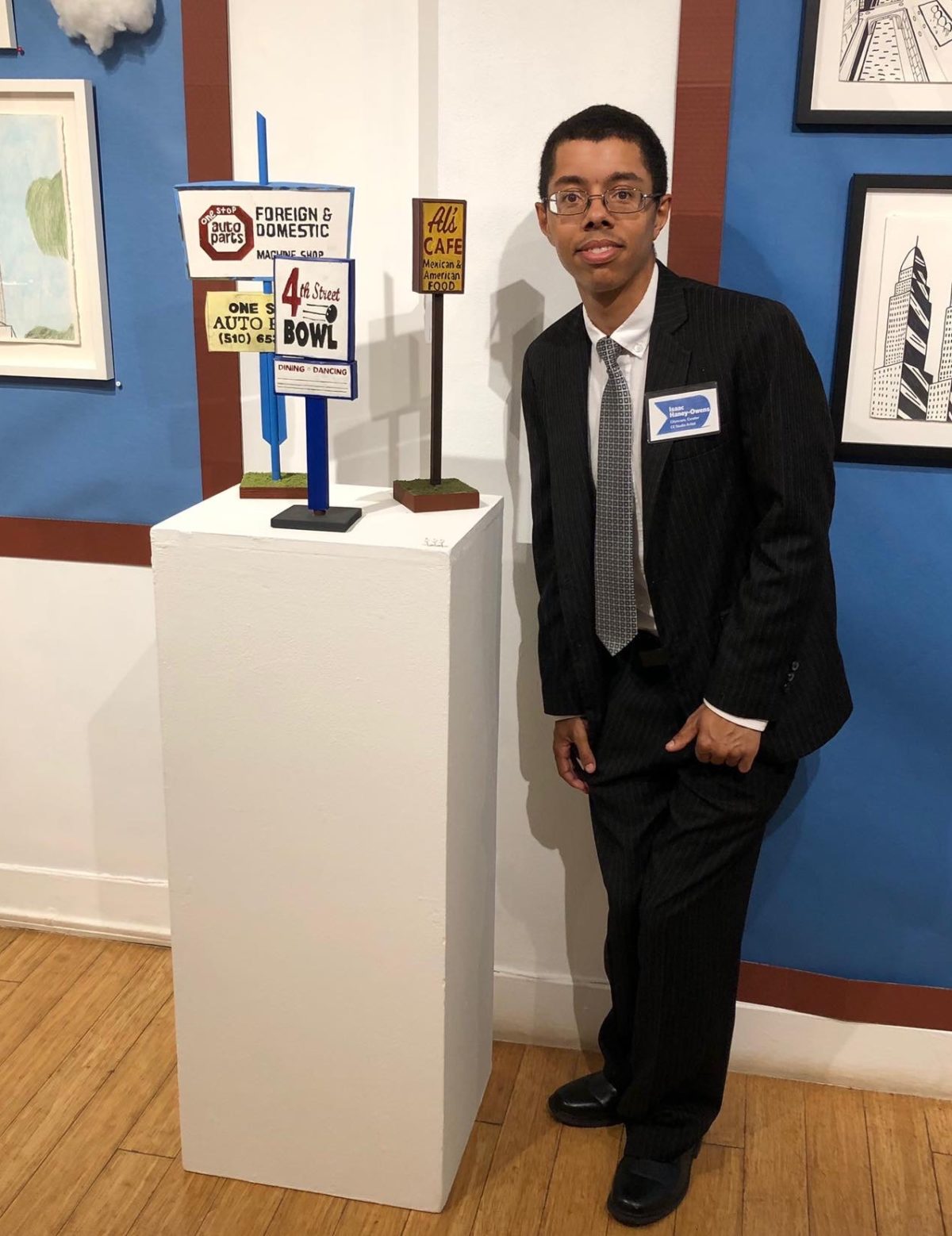 Isaac, a black young man, wearing a suit and eye glasses next to his art featured at Creativity Explored