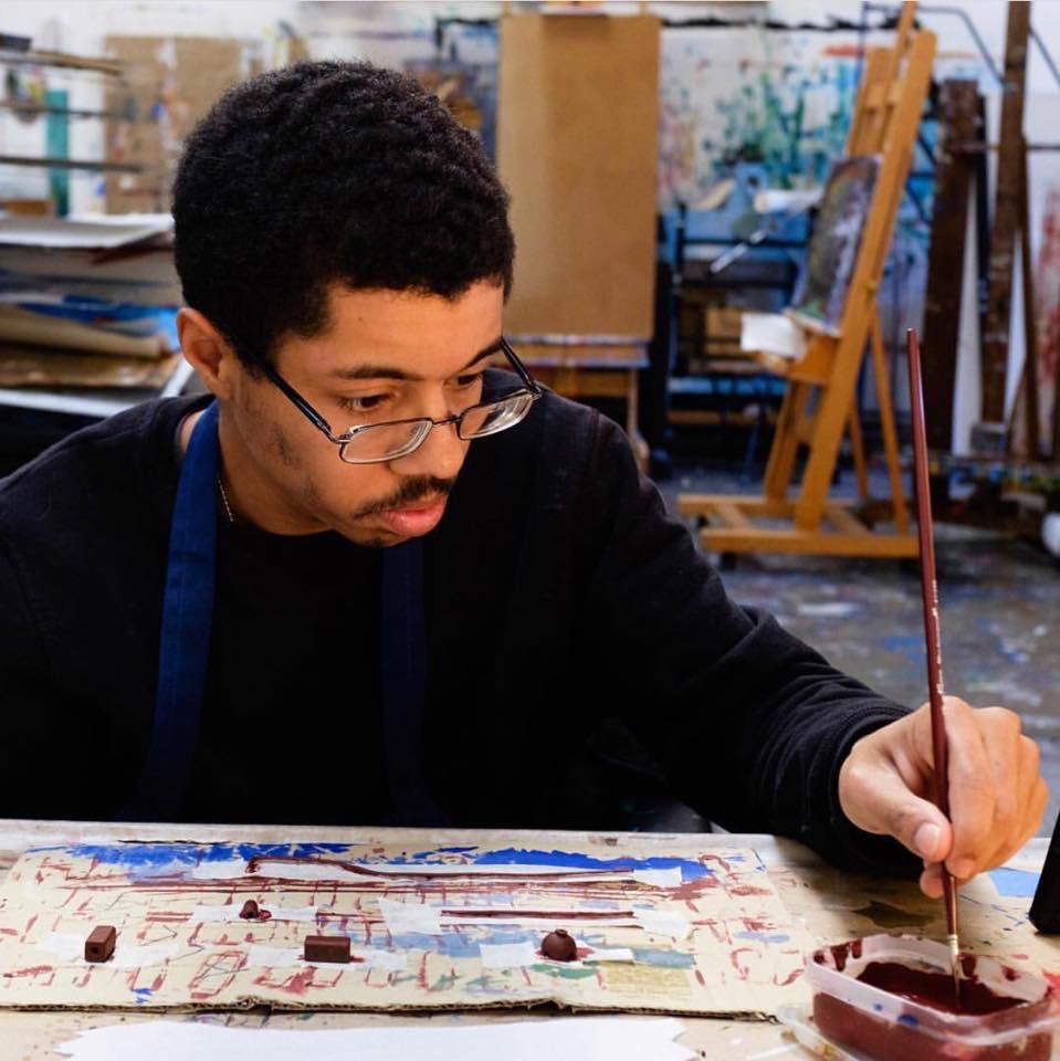 Isaac Haney Owens, a dark-skinned man with a moustache wearing glasses, is pictured painting on a piece of cardboard.