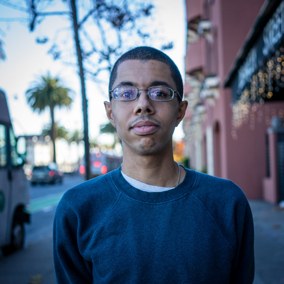 Isaac, a black young man, wearing a navy sweater and eye glasses in an urban background