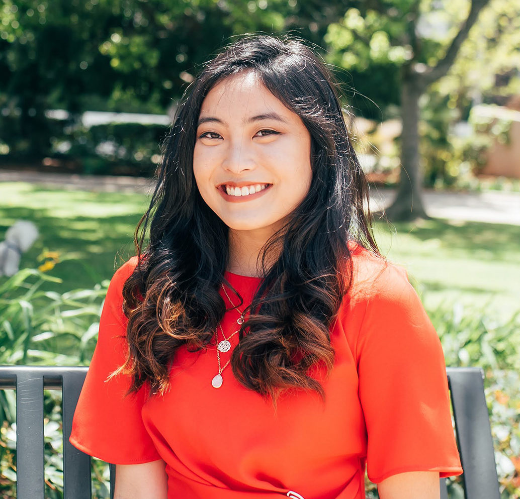 Catie, a young Asian woman in a red dress, smiles in front of a green park