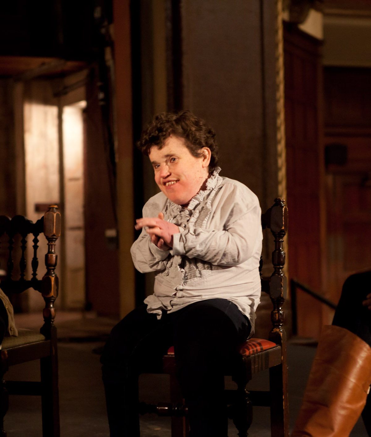 Kelsey is pictured sitting in a chair. She is a light skinned woman with short, brown hair, wearing a grayish purple shirt and dark pants. The walls in the room behind her are brown.