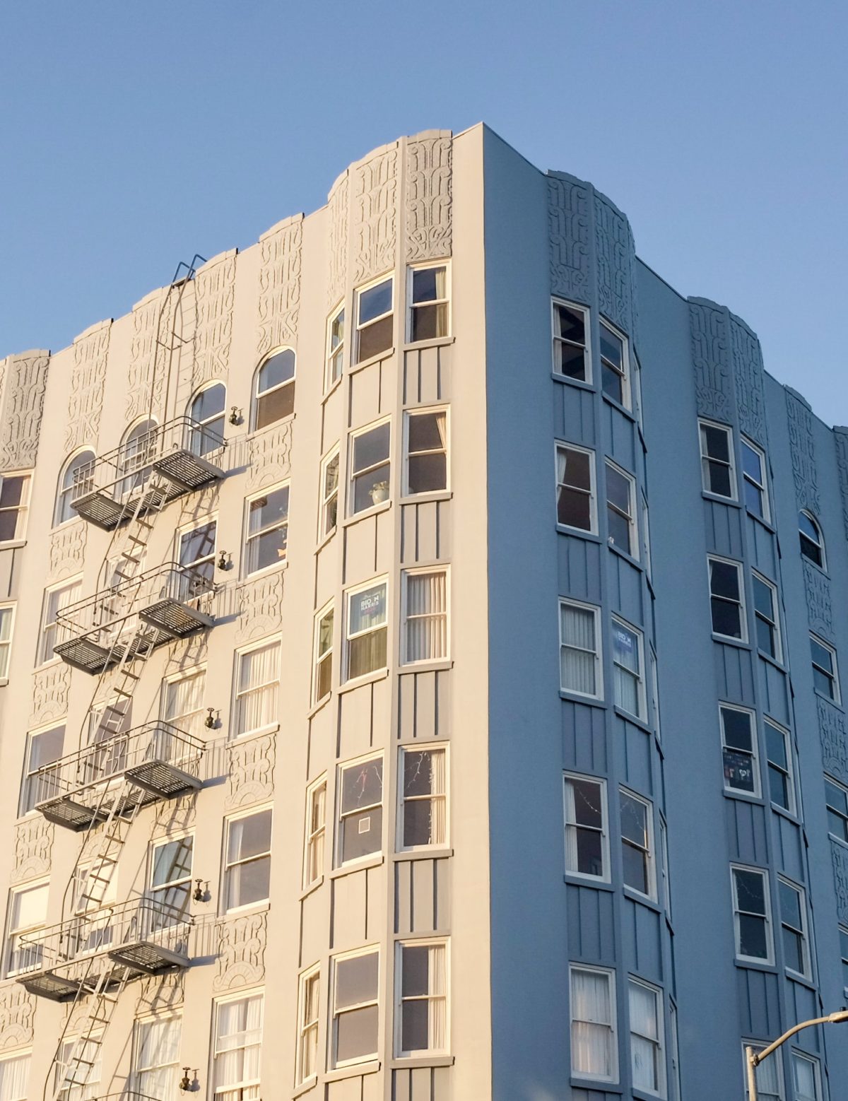 image of multifamily housing building in California. The photo looks up at the top of the building with the blue sky surrounding it. The building is white and there are 5 stories shown.