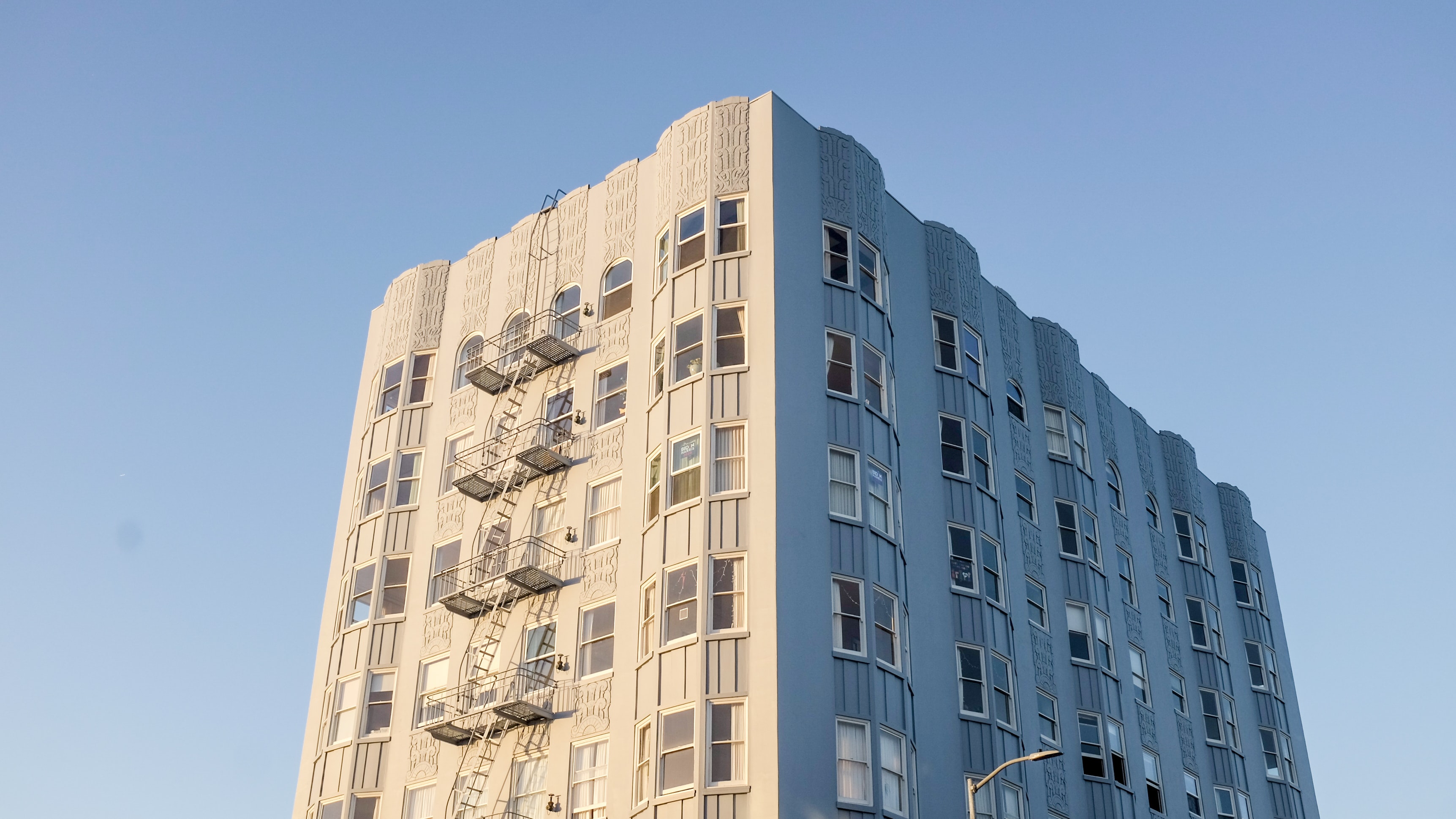 image of multifamily housing building in California. The photo looks up at the top of the building with the blue sky surrounding it. The building is white and there are 5 stories shown.