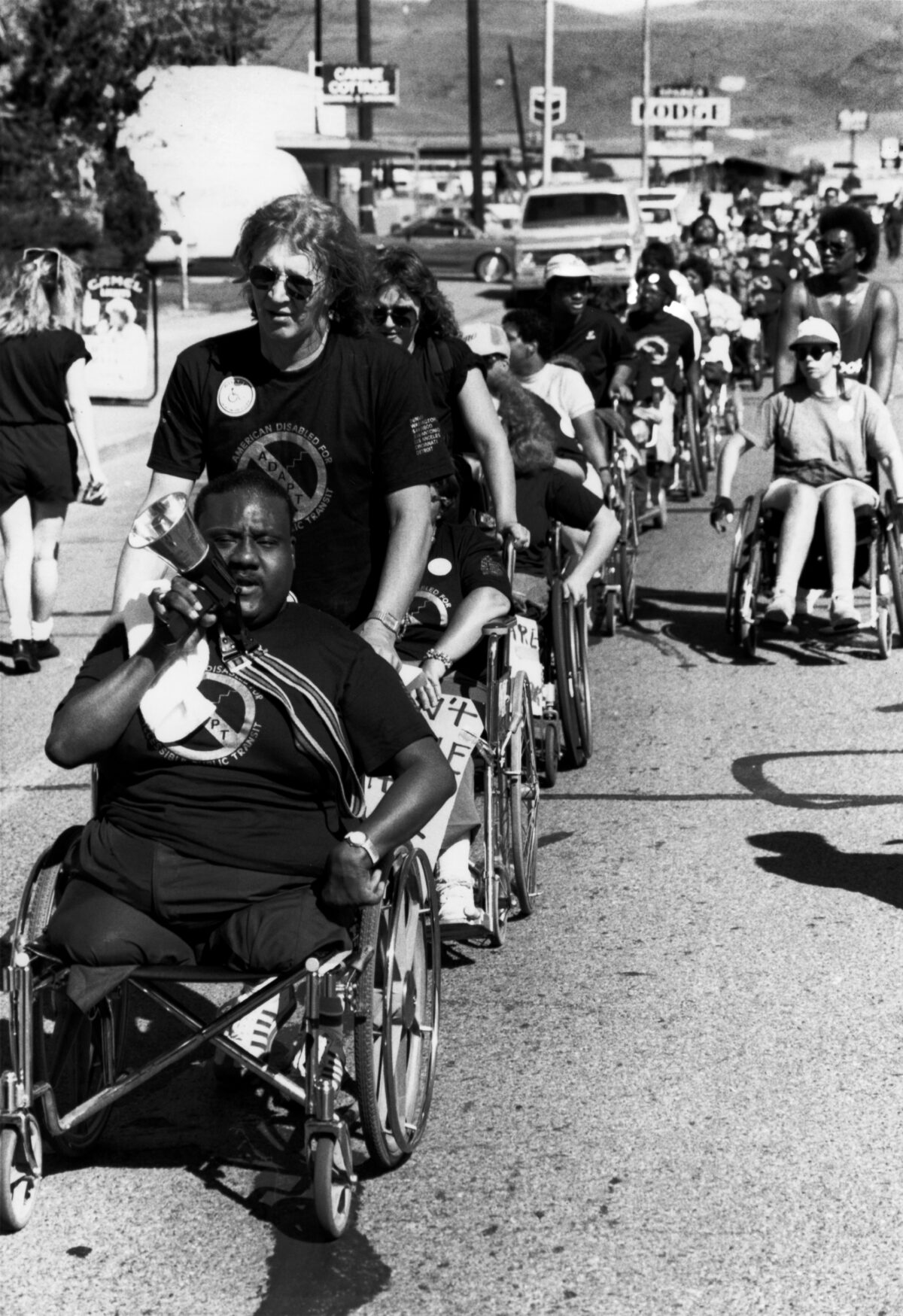 image of disability rights march with black disabled man in wheelchair at from with white man pushing him. There looks to be over 100 people present