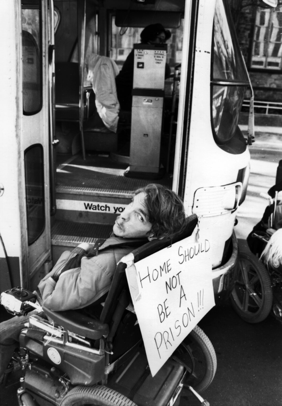 A picture of a person in a wheelchair blocking the entrance to a bus. There is a sign on the back of his wheelchair that reads "Home should not be a prison!!!"