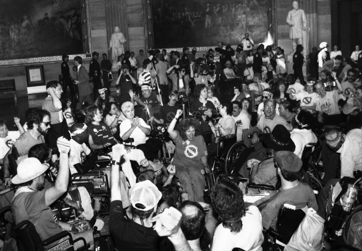 A black and white photo of a large group of protesters, some standing, many in wheelchairs, in the lobby of a building