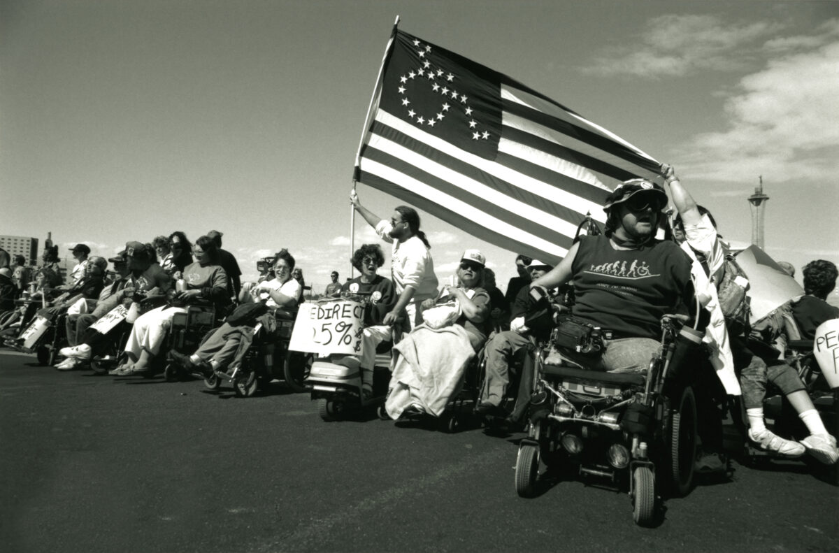 A row of people in wheelchairs with a few people standing. One person is standing holding an American flag with the stars on it in the shape of someone sitting in a wheelchair.