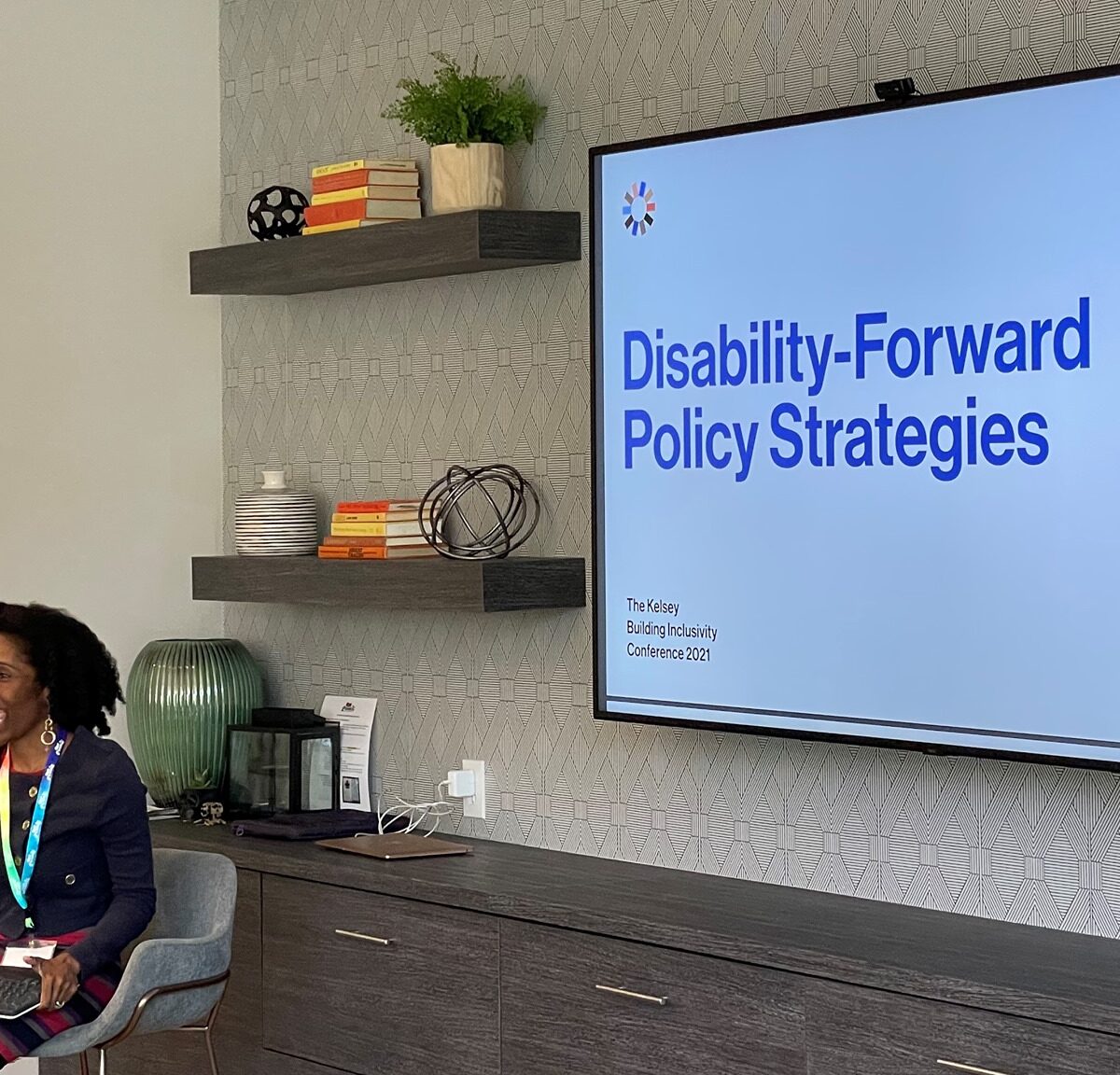 Photograph of a black woman with curly hair sitting in a chair in front of a slide on a screen that reads "Disability-Forward Policy Strategies"