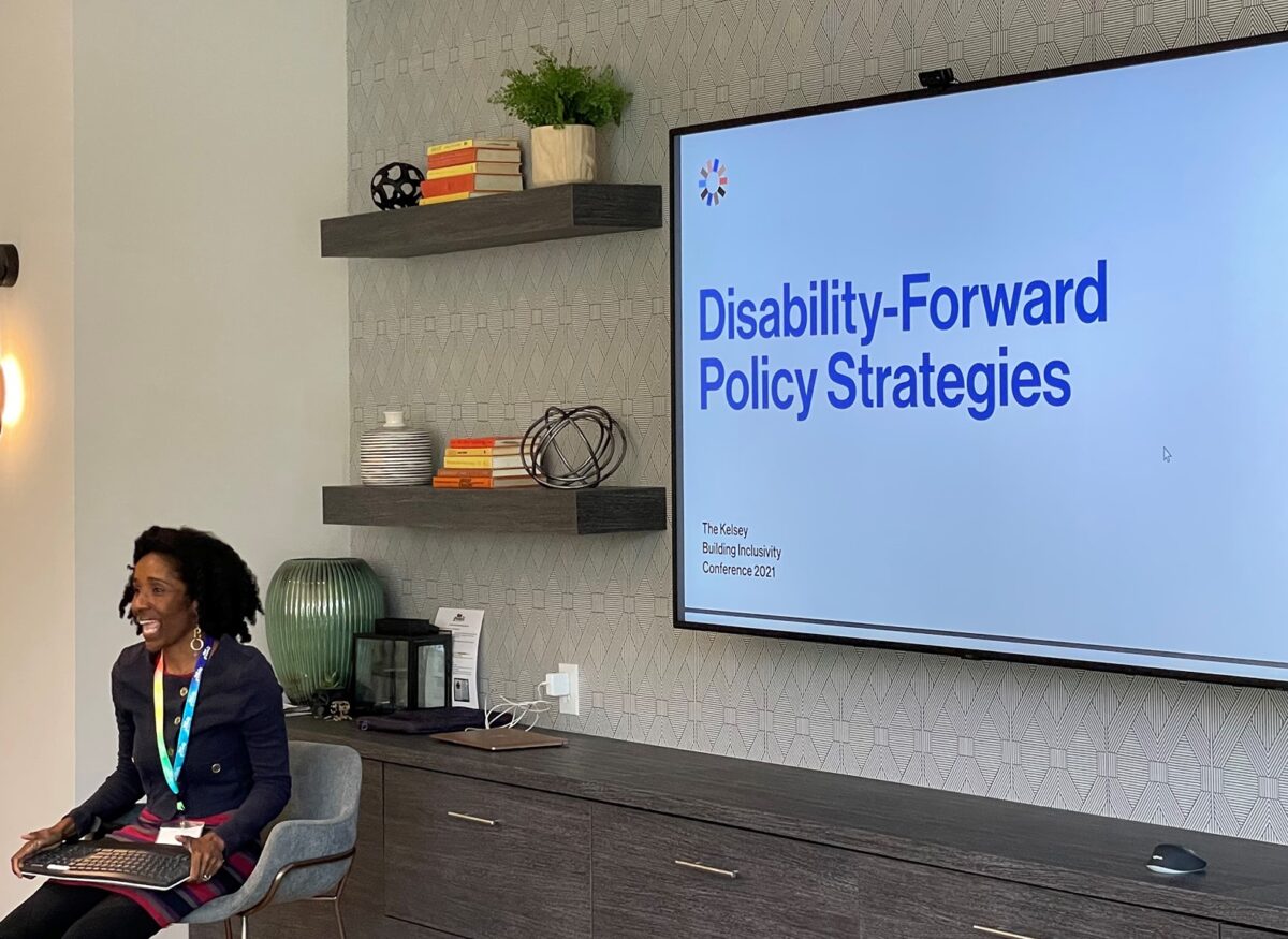 Photograph of a black woman with curly hair sitting in a chair in front of a slide on a screen that reads "Disability-Forward Policy Strategies"