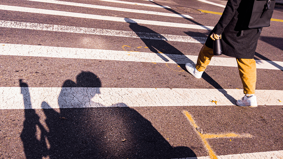 The shadow of a person in a wheelchair can be seen in the crosswalk of a street. The legs of a person crossing the crosswalk are pictured.