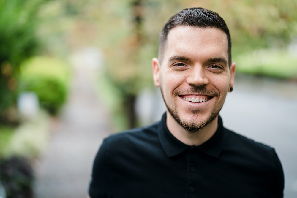 Image Description: Against a blurred background with greenery, Dom, a light-skinned man in a black shirt, is pictured smiling.