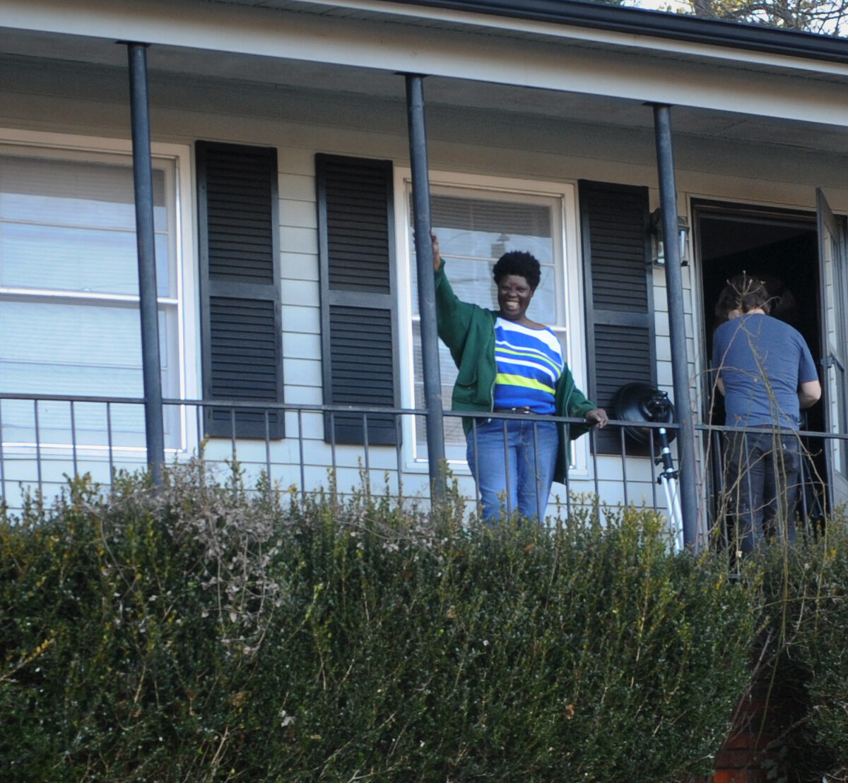 Photo by Tom Olin of Lois Curis, Black disabled woman and plaintiff from the US Supreme Court Case of Olmstead, smiling waving to people facing her. She is standing in front of her own home.