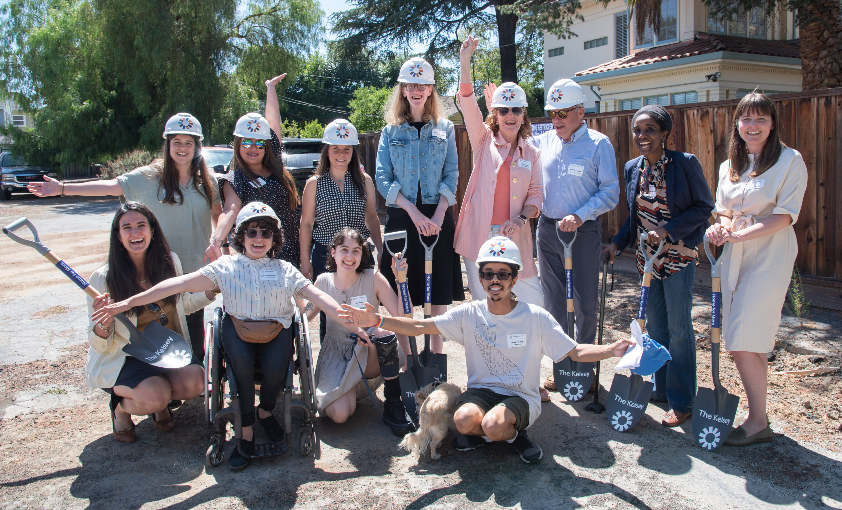 A photo of The Kelsey team and Kelsey's mom and dad, including 11 people with and without visible and invisible disabilities, of difference races and ages. Some people wearing white hard hats with The Kelsey logo and others are holding shovels.