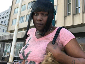 Lisa, a dark-skinned woman, is wearing a pink t-shirt with dark straight hair and bangs. She is standing in front of a concrete building.