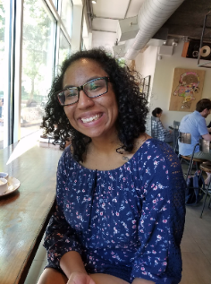 Biracial person with curly black hair is smiling for the camera. She has black-rimmed glasses and is wearing a blue, flowered romper. Valerie is seated in a coffee shop, backs of patrons visible behind her.