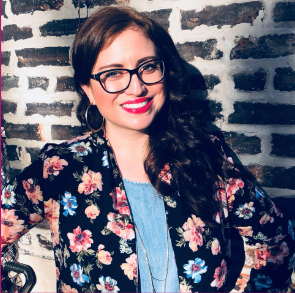 Katherine, a cis-gendered light-skinned Latina woman with long brown wavy hair and glasses, smiling at the camera.