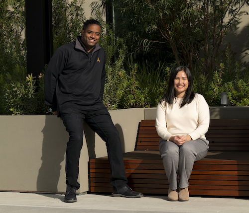 A young man and a woman sit with big smiles in an outdoor space with beautiful greenery behind them. One woman is African American, dressed in all black. The other young woman is Asian with a white top and grey pants. See less