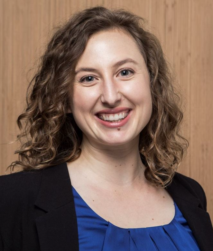 Headshot of a middle aged white woman smiling with curly hair, wearing a navy blue under a black blazer. She stands in front of a wood colored background.