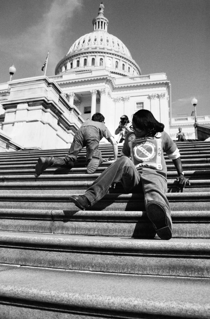 Black and white photo of capitol crawl, in which two people, one dark shinned and one light skinned are crawling up the US capital steps. Photo taken during the fight for the passage of the Americans with Disabilities Act. 