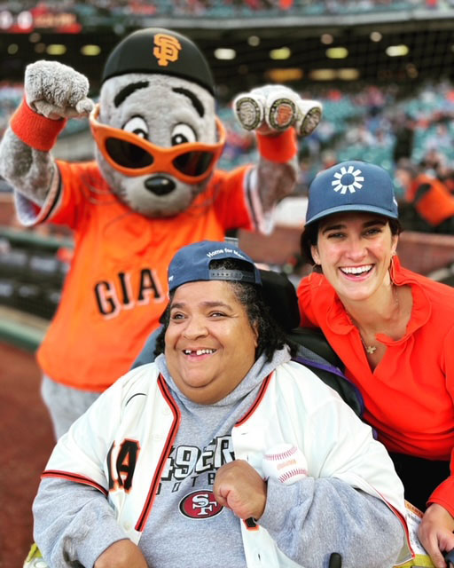 Liz, a black woman in a wheelchair wearing a hat and baseball Jersey over a sweater, stands with the Giants mascot, and The Kelsey Co-Founder Micaela Connery. Micaela is a white woman with dark hair.