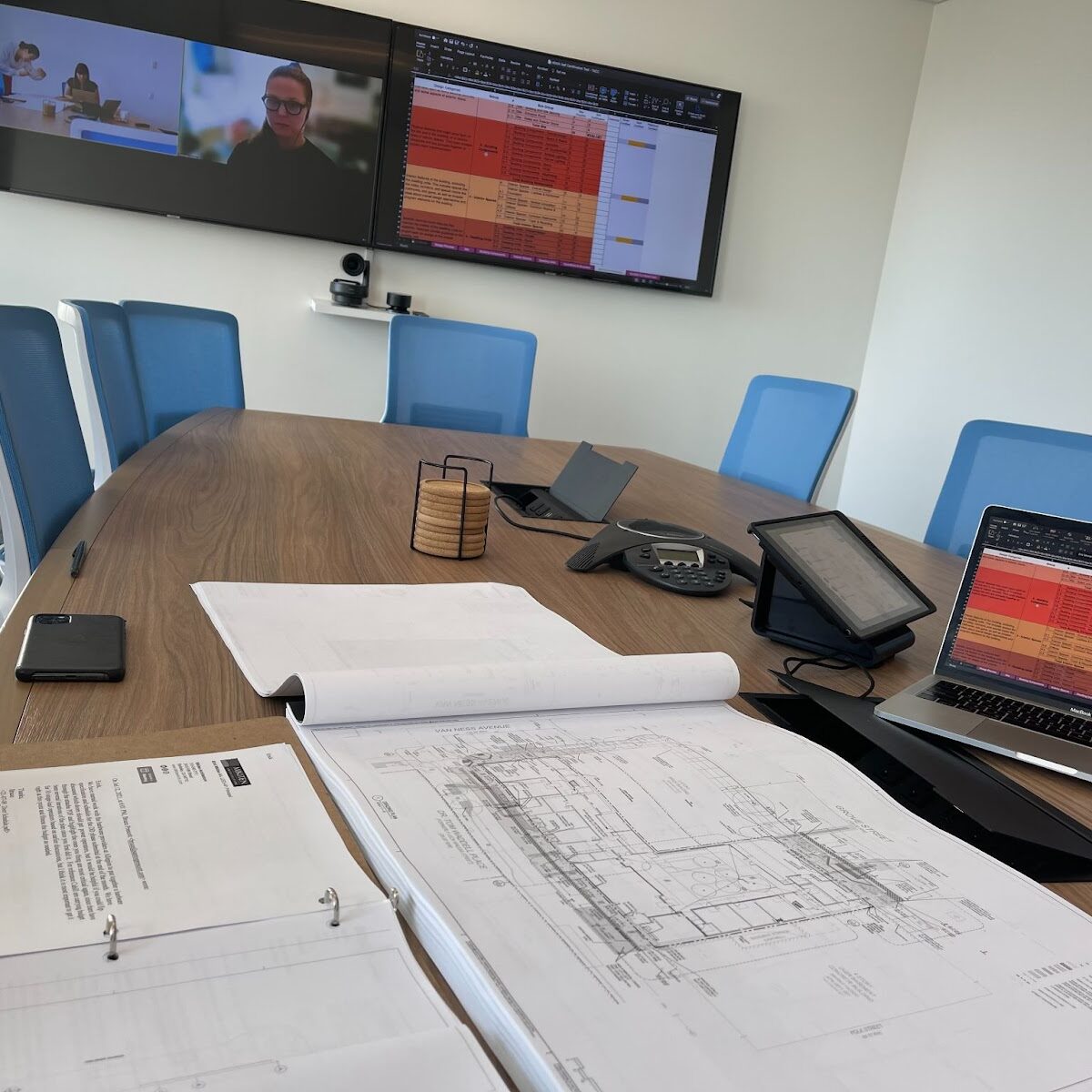 A conference room table is seen with two screens on the wall behind it. There are schematic drawings, notebooks, a laptop, coasters and an audio conference phone is seen on the table.