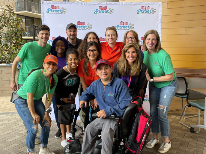 Happy group shot with 12 people in 2 rows with brightly colored t-shirts including green, red and purple. Man in a power wheelchair is central.