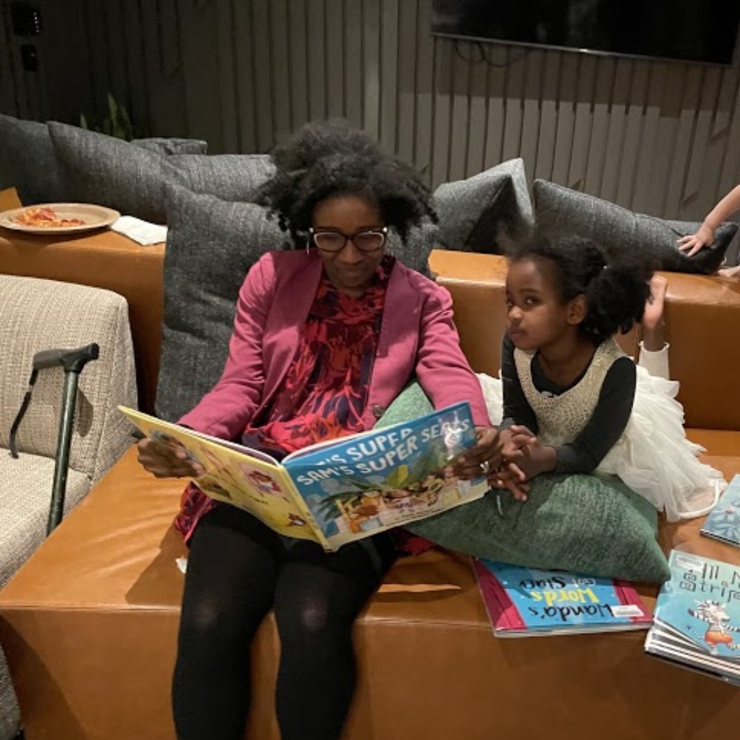 A dark-skinned woman reads a book to a dark-skinned girl. They are sitting on a brown leather seat and there are cushions behind them.