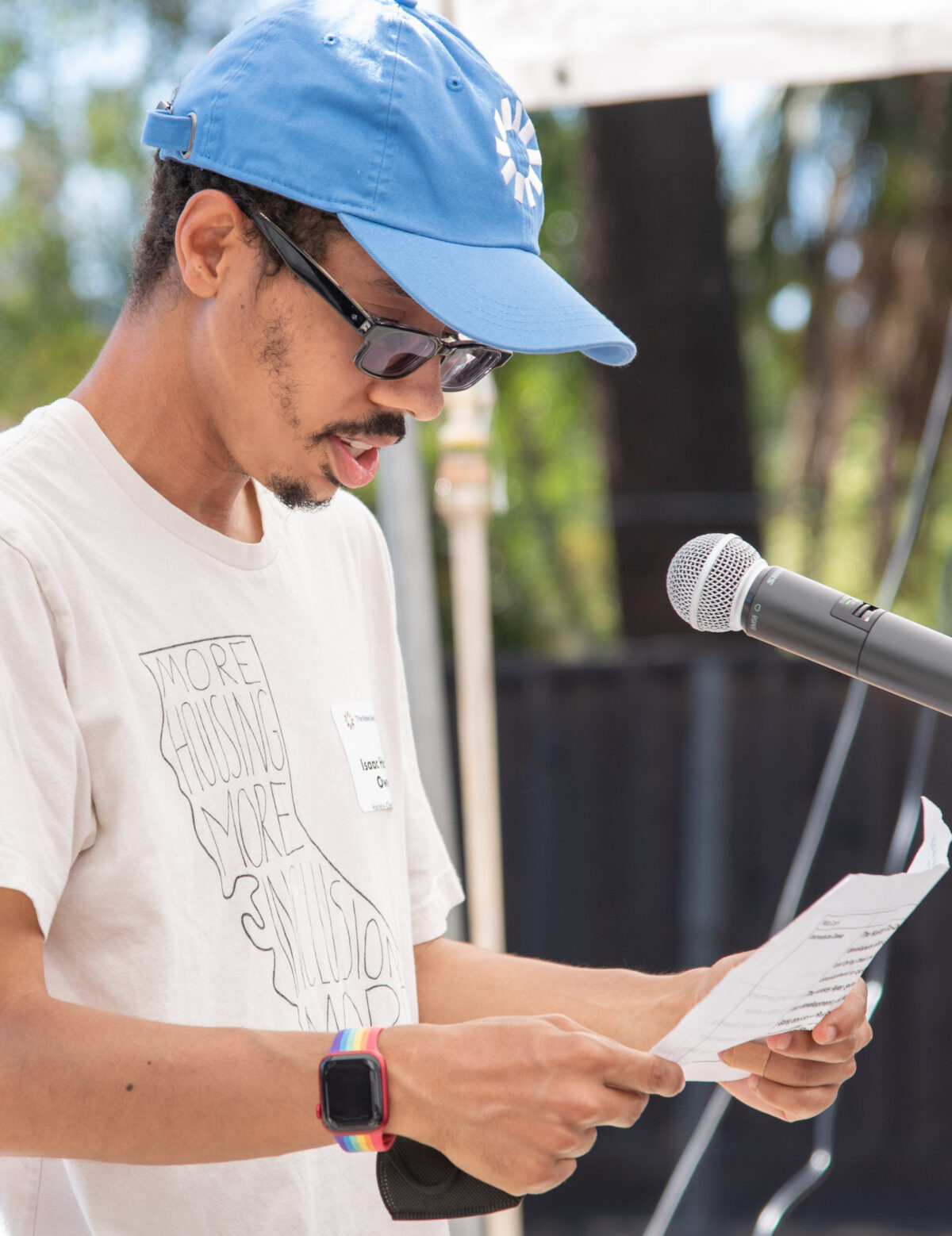 Isaac Haney Owens, a dark-skinned man with a beard wearing glasses, holds a paper and stands speaking behind a microphone