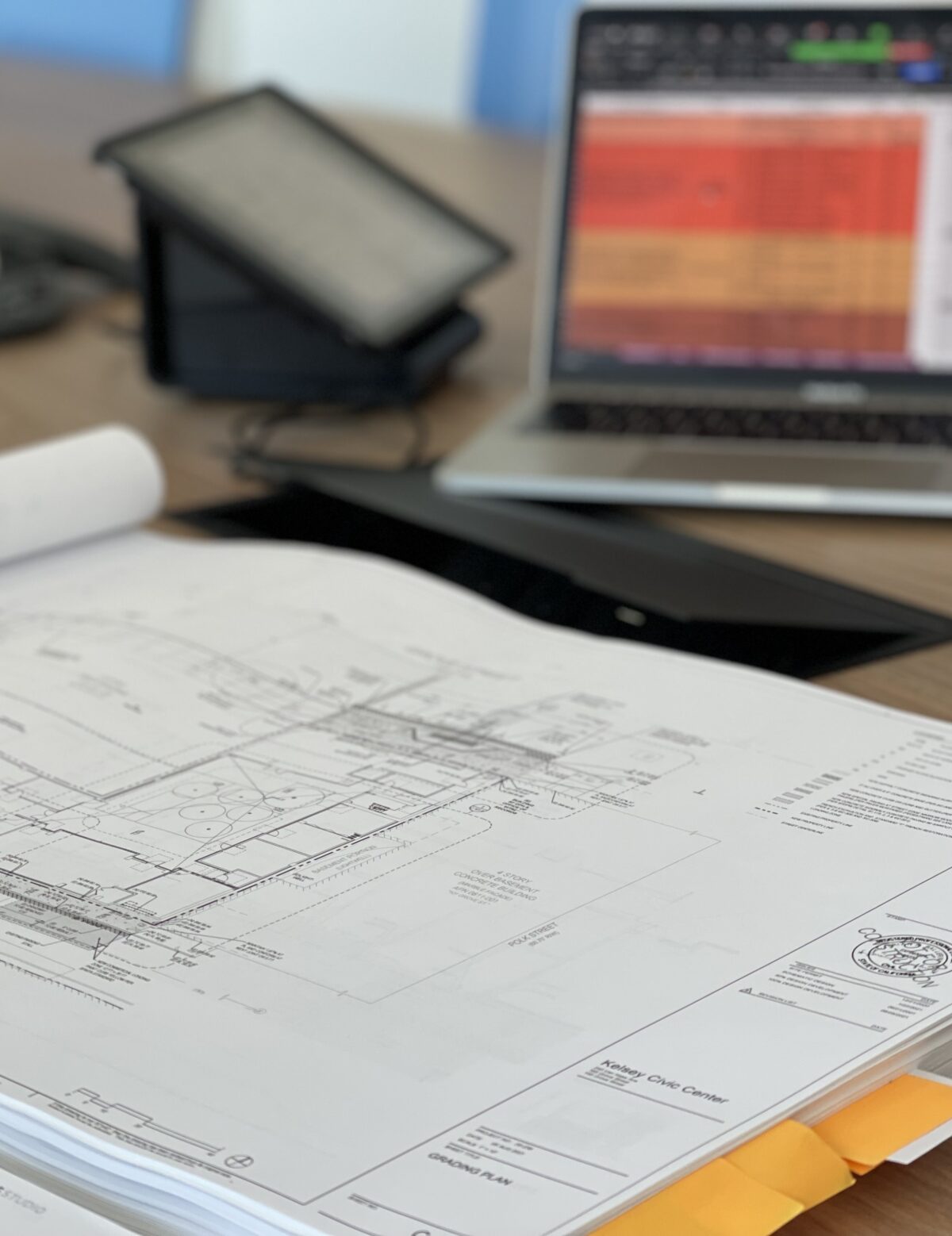 Schematic drawings lie on a table. A tablet and a laptop can be seen in the background