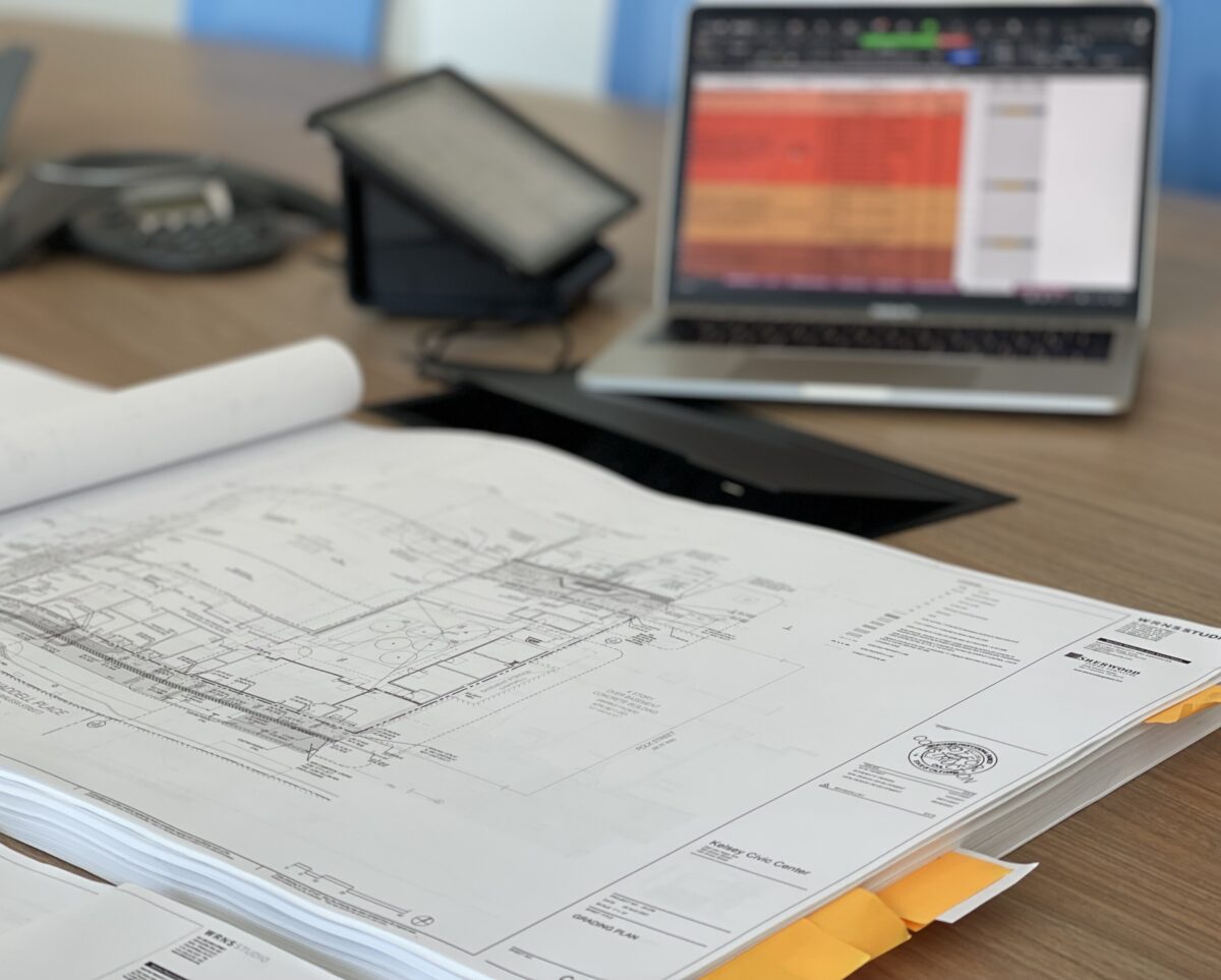 Schematic drawings lie on a table. A tablet and a laptop can be seen in the background