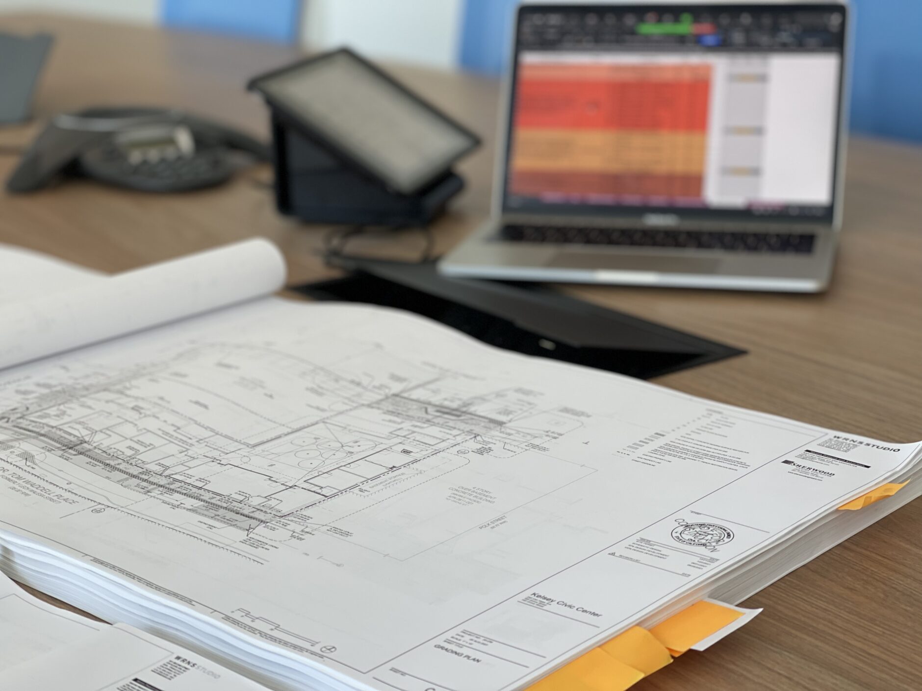 Schematic drawings lie on a table. A tablet and a laptop can be seen in the background