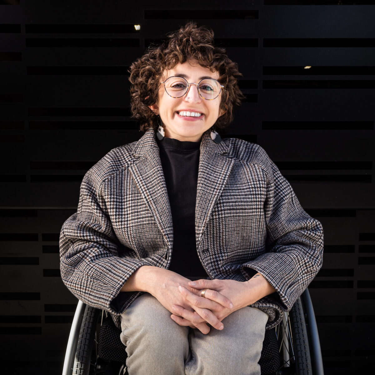 White queer person smiling in their manual wheelchair with short brown hair, round glasses, earrings, and a blazer.